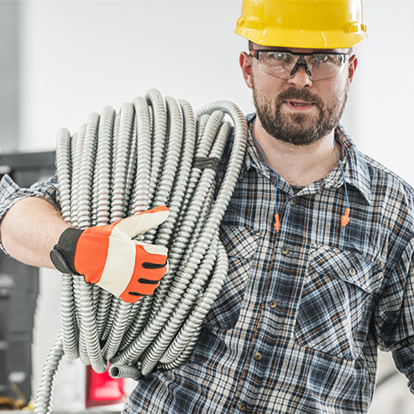Hombre con casco amarillo y gafas protectoras, vistiendo camisa de cuadros, cargando una gran bobina de tubo flexible metálico en su hombro y usando guantes de seguridad naranjas y blancos.