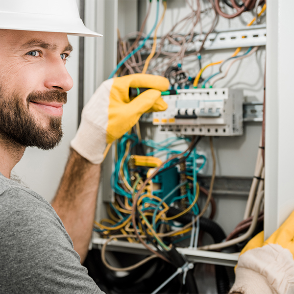 Electricista sonriente con casco blanco y guantes amarillos, ajustando un interruptor en un panel eléctrico lleno de cables de colores.