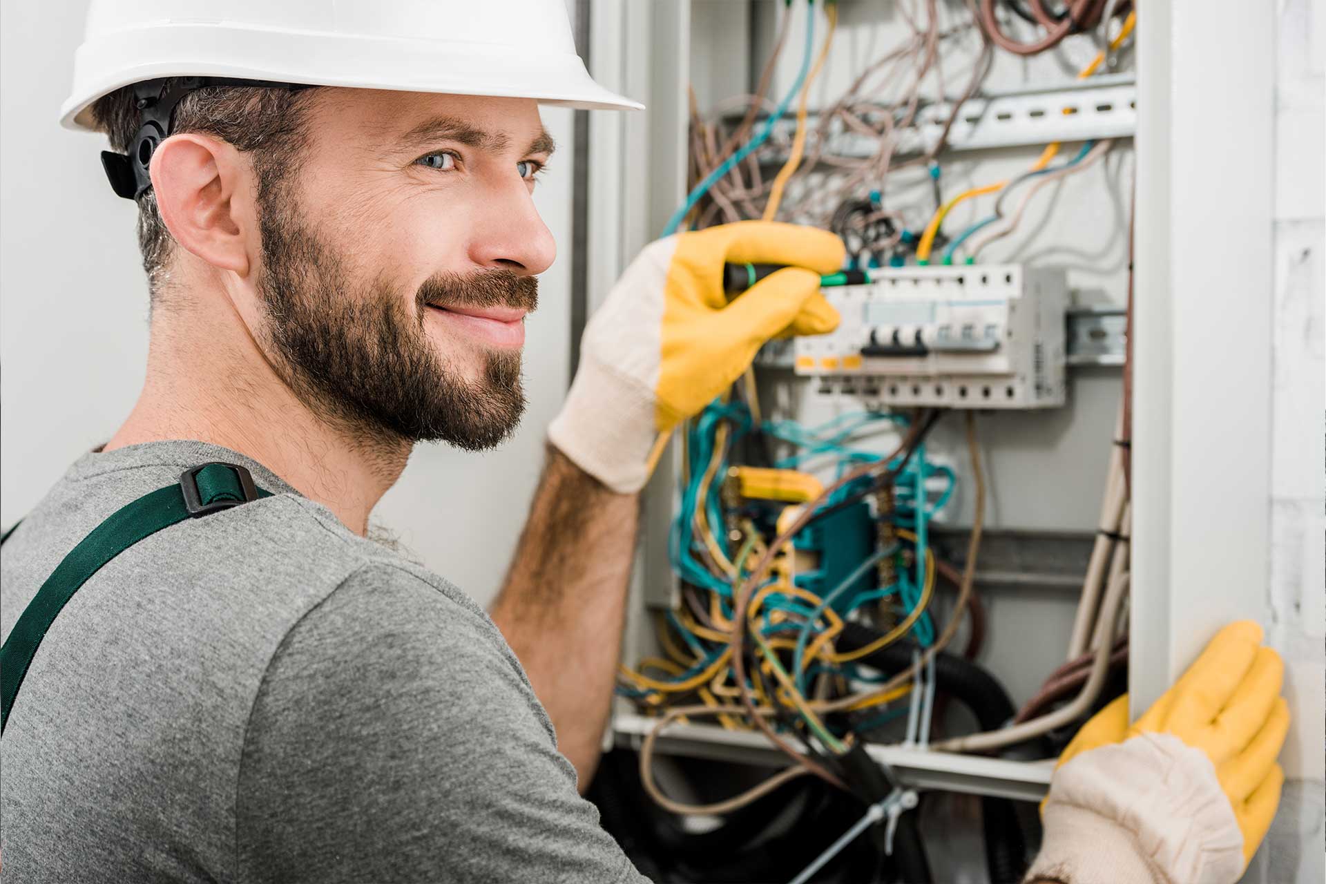 Electricista sonriente con casco blanco y guantes amarillos, ajustando un interruptor en un panel eléctrico lleno de cables de colores.