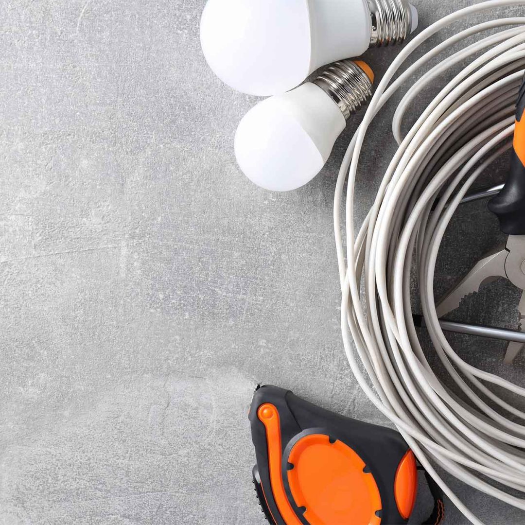 Bobinas de cable blanco junto a herramientas como pinzas y destornilladores con mangos naranjas, además de dos bombillas y una cinta métrica.